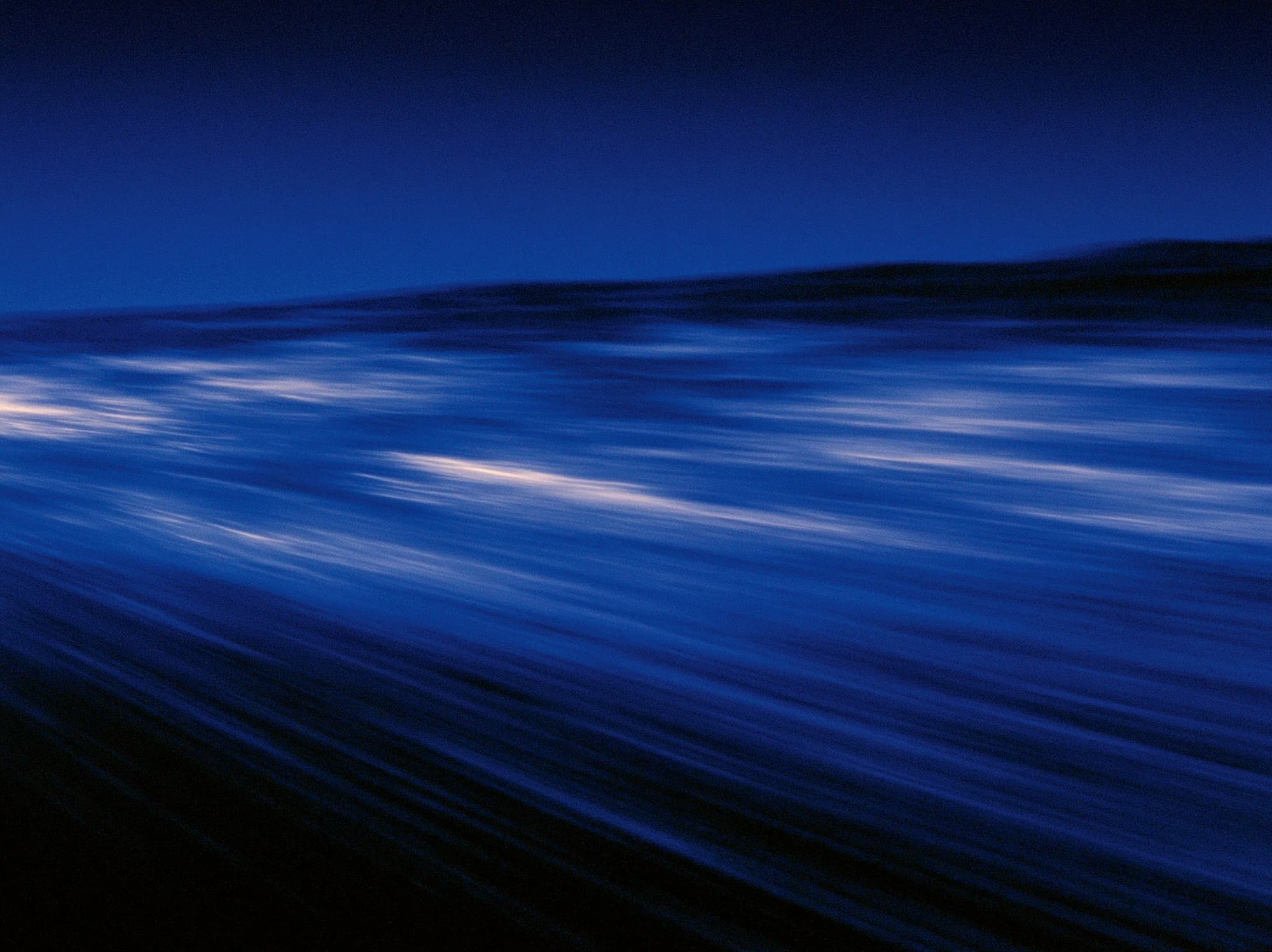 Verwischtes dunkelblaues Wasser in der Dämmerung, helle Lichtstreifen auf den Wellen und ein flacher Hügelzug am Horizont.
