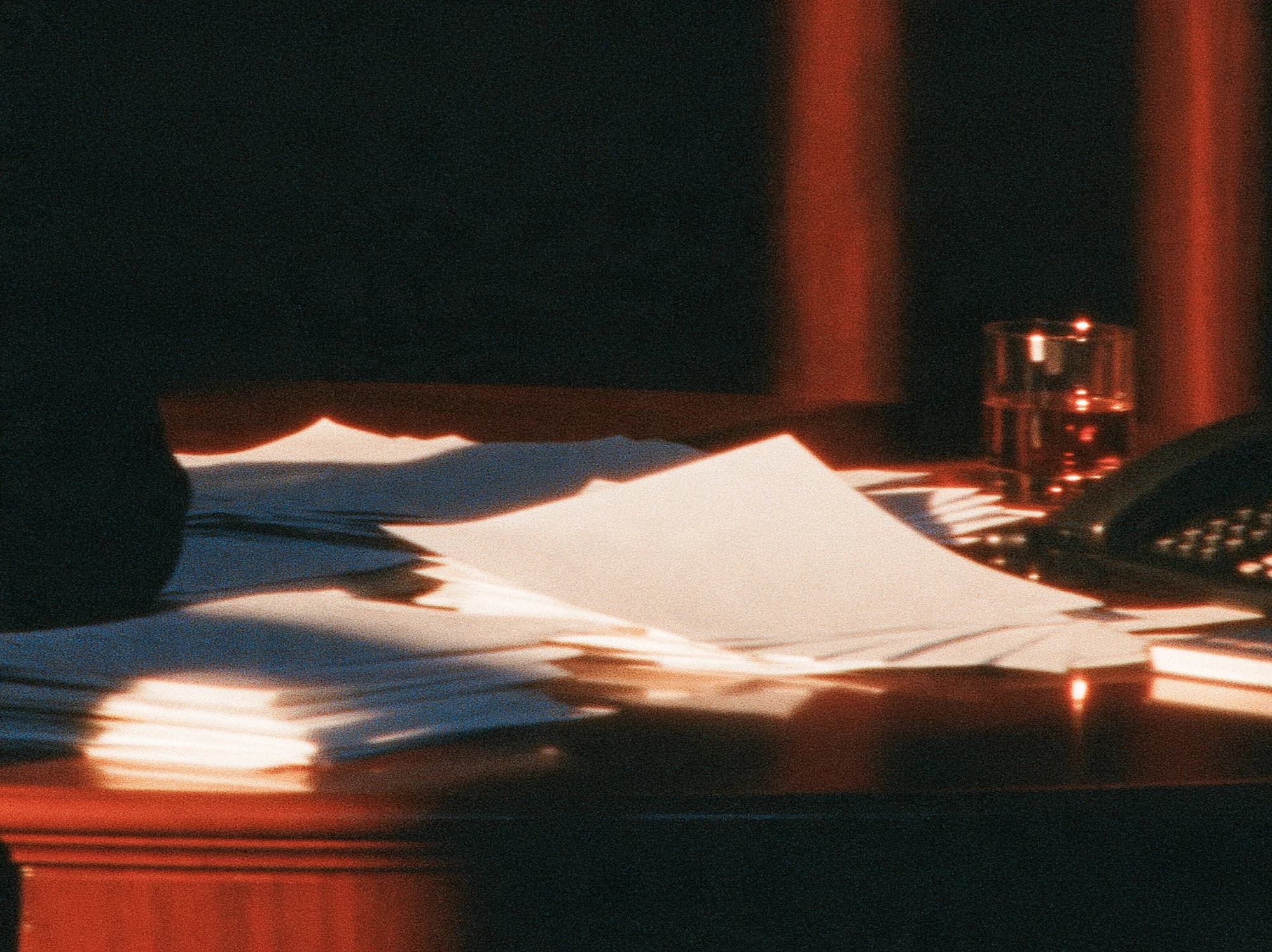 Papiere auf einem dunklen Schreibtisch im orangefarbenen Licht, neben einem Glas und einem Telefon.